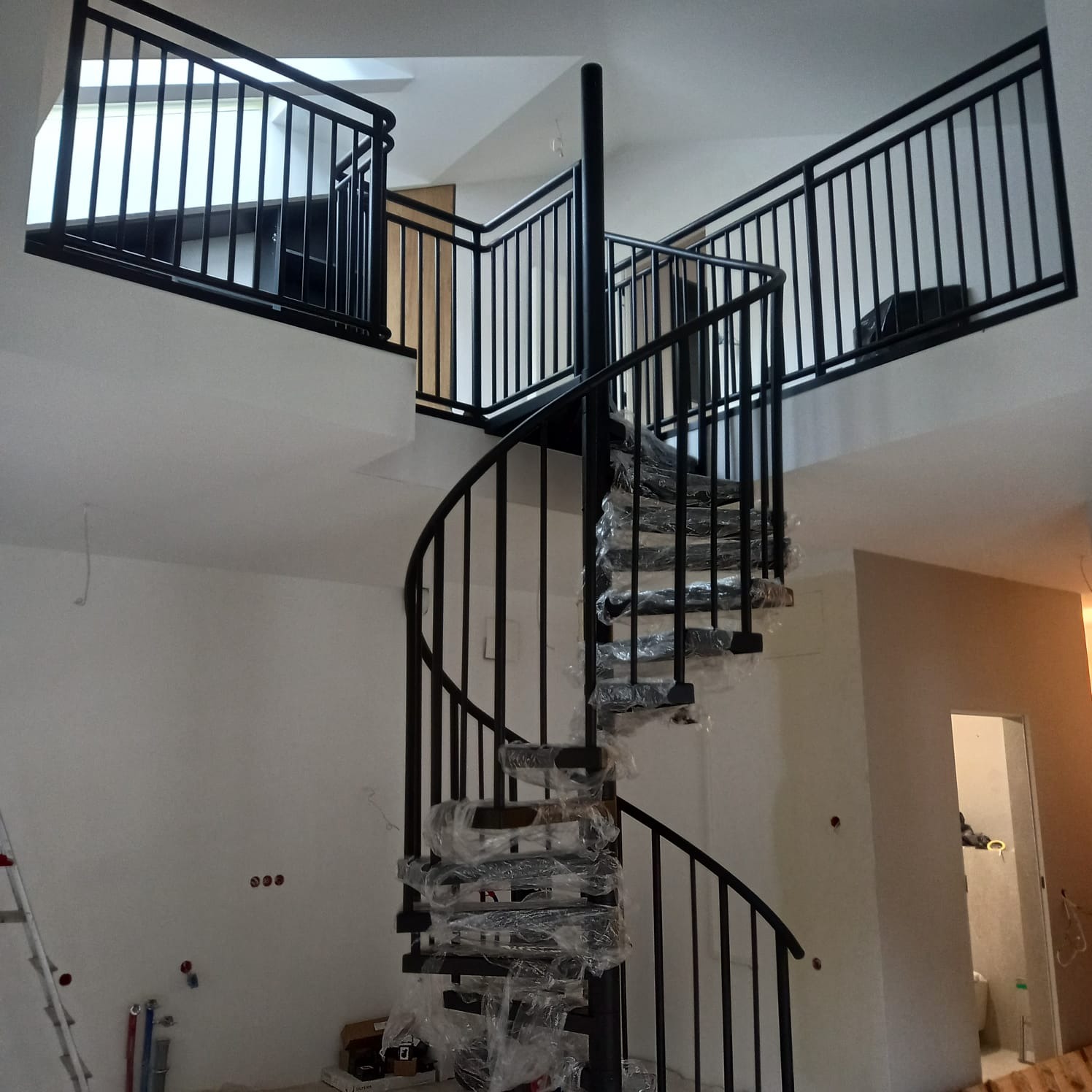 Czarna, spiralna konstrukcja schodowa z barierkami na dwóch poziomach, jeszcze w folii ochronnej, w trakcie montażu w surowym wnętrzu.