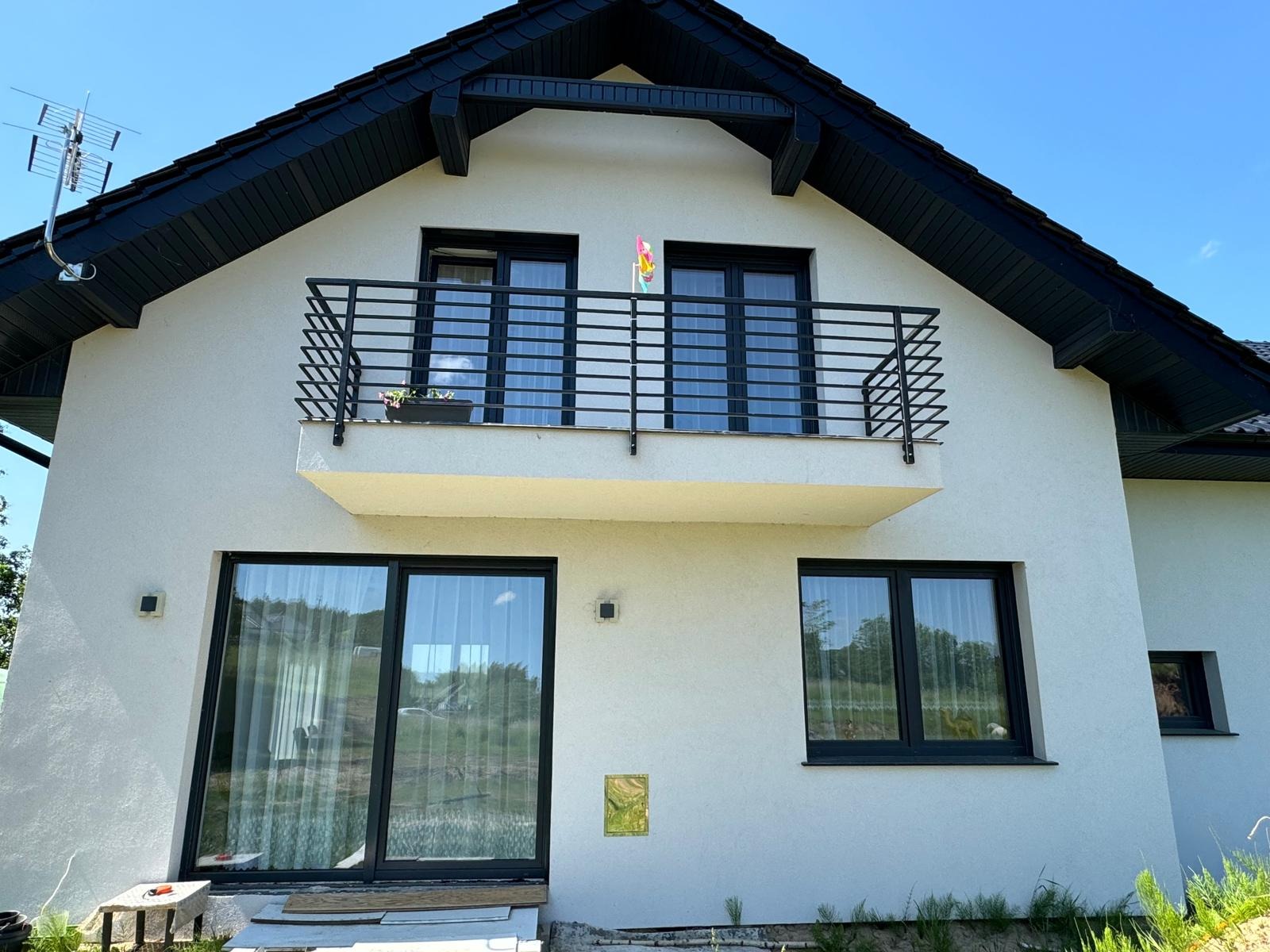 Biały dom z czarnym dachem i czarną, metalową balustradą balkonową o prostym wzorze, z kwiatami w doniczce; widoczne duże okna i drzwi balkonowe z czarnymi ramami.