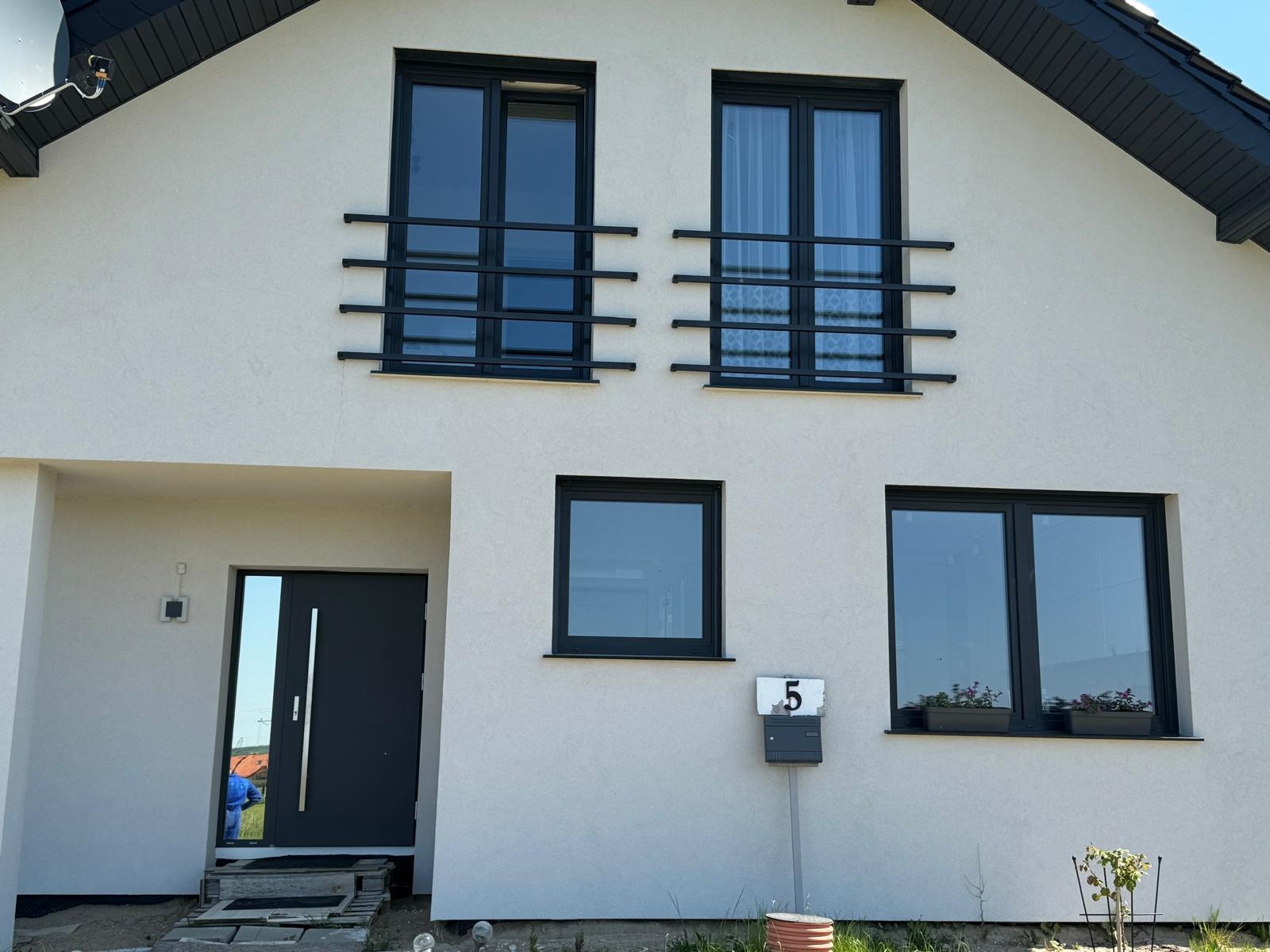 Fasada domu jednorodzinnego z czarnymi oknami zabezpieczonymi prostymi, poziomymi barierami oraz ciemnoszarymi drzwiami wejściowymi z długim, pionowym uchwytem, widoczna skrzynka na listy z numerem 5.