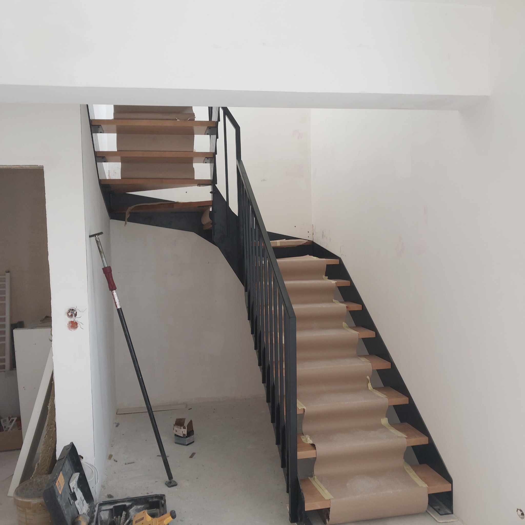 Czarne schody metalowe z drewnianymi stopniami, zabezpieczone papierem, w jasnym wnętrzu w trakcie wykończenia. Widoczna podpora teleskopowa i narzędzia.