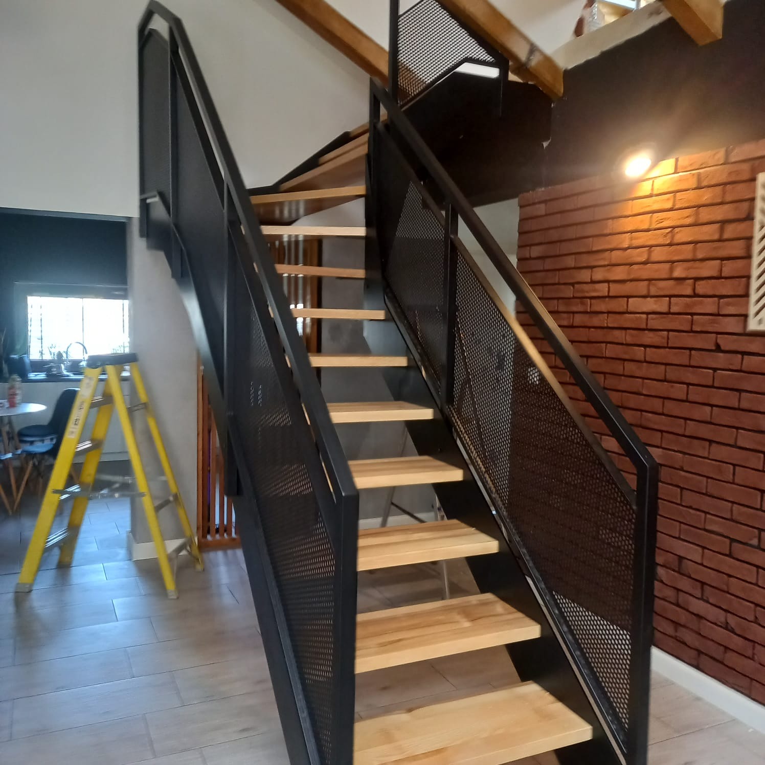 Nowoczesne schody z drewnianymi stopniami i czarną, perforowaną metalową balustradą w industrialnym wnętrzu z ceglaną ścianą i żółtą drabiną z boku.