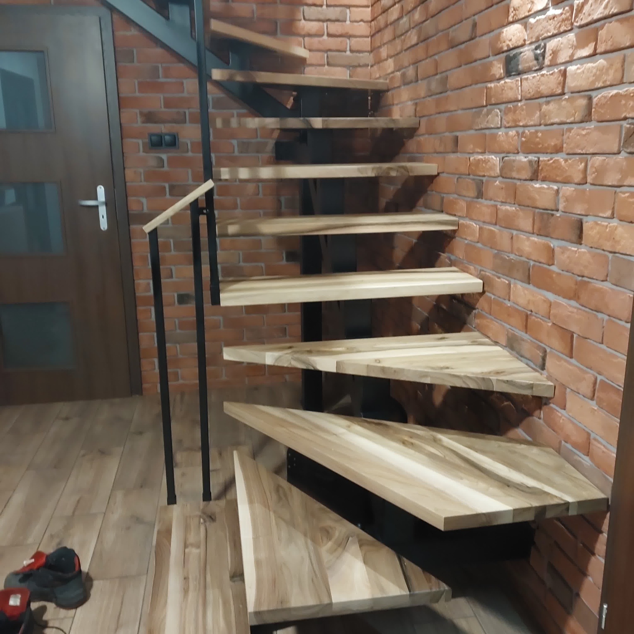 Drewniane schody wspornikowe z czarną metalową konstrukcją, zamontowane przy ceglanej ścianie. Widoczne drewniane stopnie i fragment poręczy. Nowoczesny design.