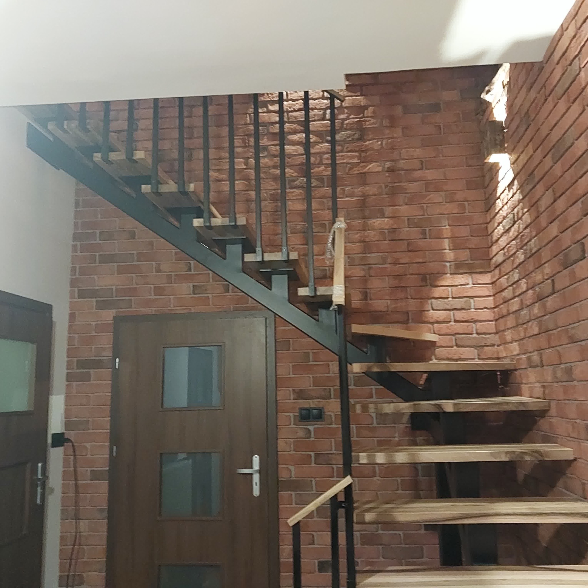 Metalowe schody z drewnianymi stopniami na tle ceglanej ściany. Nowoczesny design z czarną konstrukcją i jasnym drewnem. Widok z dołu, częściowo widoczne drzwi.