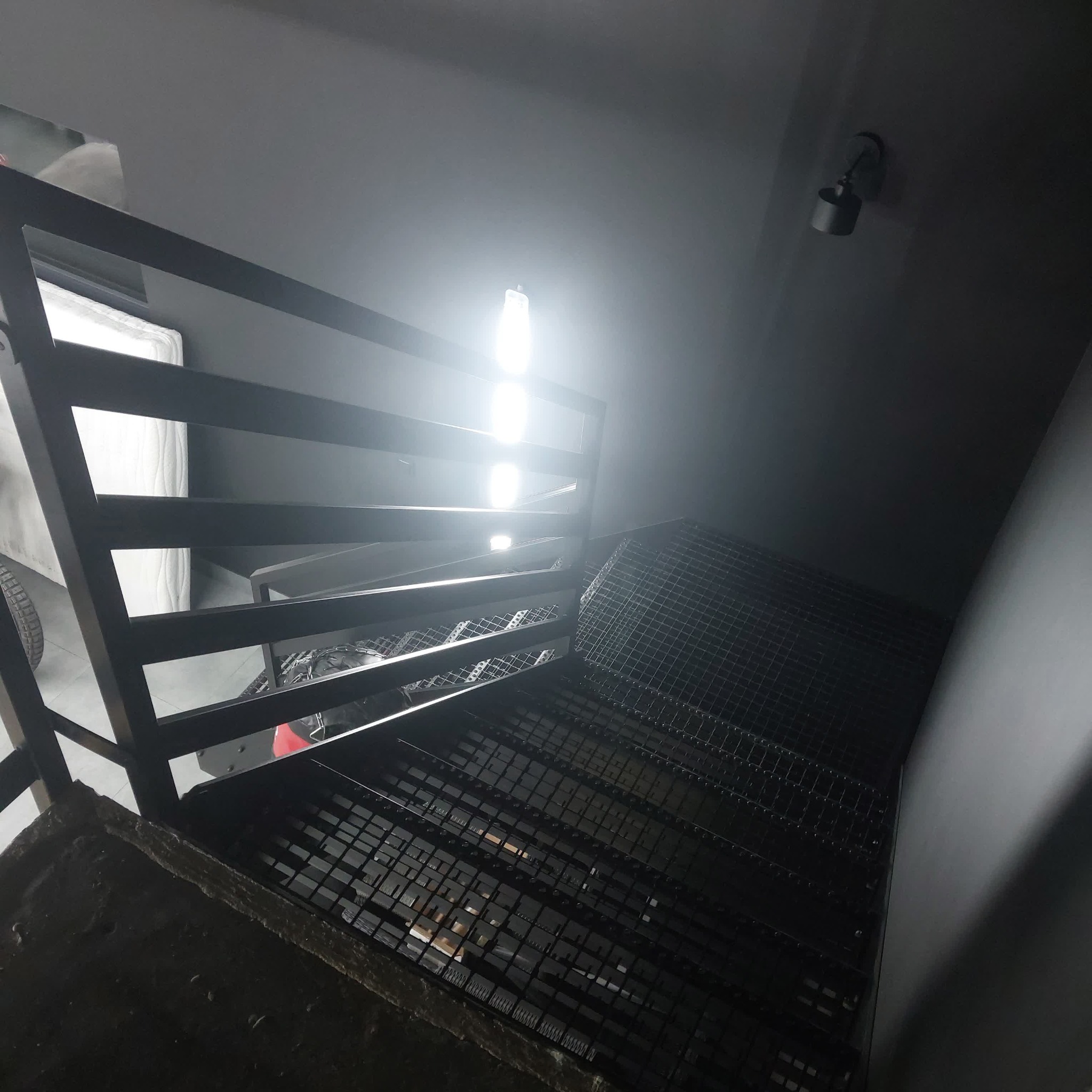 Nowoczesne schody z metalową kratą i minimalistyczną balustradą w ciemnej tonacji, oświetlone punktowo, widziane z dołu pod kątem, z fragmentem opony w tle.
