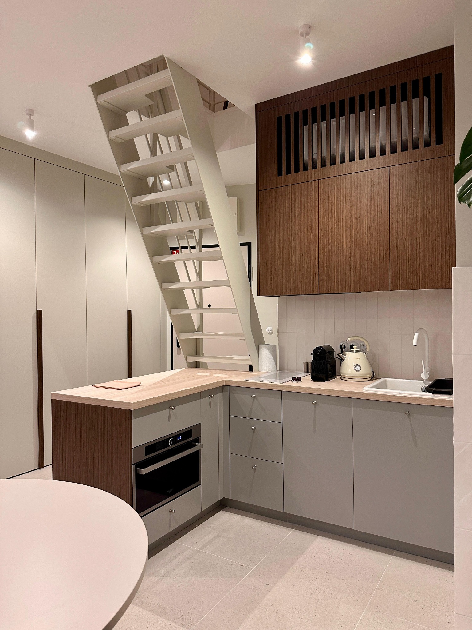 Nowoczesna kuchnia z białymi schodami metalowymi prowadzącymi na górę, drewniane akcenty i szare szafki. Minimalistyczny design z dbałością o detale.