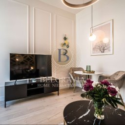 Belgravia Home Spółka z o.o. - Eleganckie wnętrze salonu z telewizorem na czarnej szafce RTV, okrągłym stolikiem kawowym z marmurowym blatem i bukietem ciemnoróżowych piwonii, okrągłym stolikiem jadalnym z dwoma krzesłami...