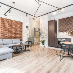 Belgravia Home Spółka z o.o. - Jasne, przestronne wnętrze mieszkania z ceglaną ścianą, szarą sofą narożną, drewnianą podłogą, ciemnymi meblami kuchennymi z wzorem w jodełkę i okrągłym stołem z czarnymi krzesłami.