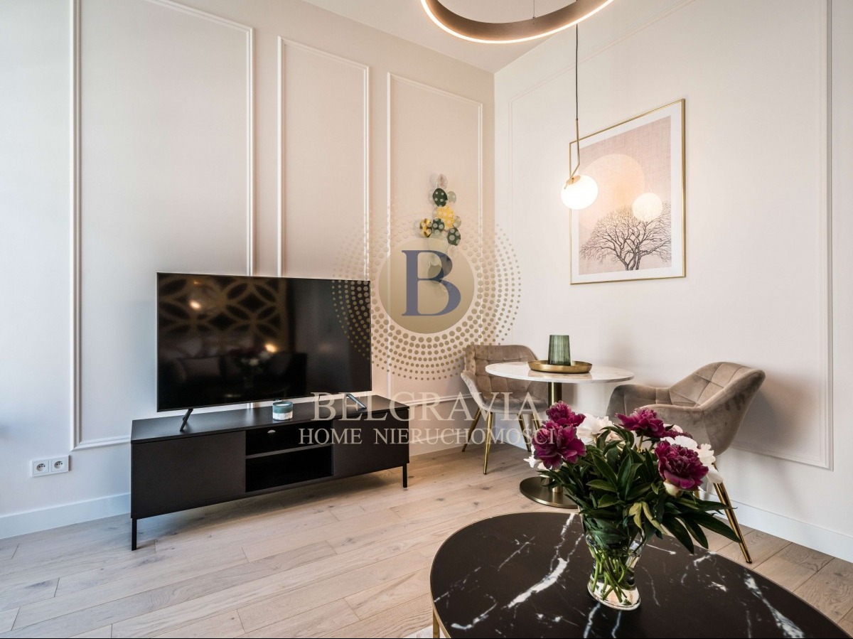 Eleganckie wnętrze salonu z telewizorem na czarnej szafce RTV, okrągłym stolikiem kawowym z marmurowym blatem i bukietem ciemnoróżowych piwonii, okrągłym stolikiem jadalnym z dwoma krzesłami...