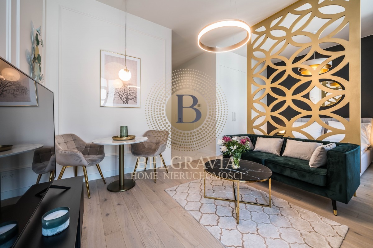 Nowoczesne wnętrze mieszkania z zieloną sofą, marmurowym stolikiem kawowym, okrągłą lampą sufitową i dekoracyjną ścianką działową w kolorze złotym, z widocznym łóżkiem w tle.