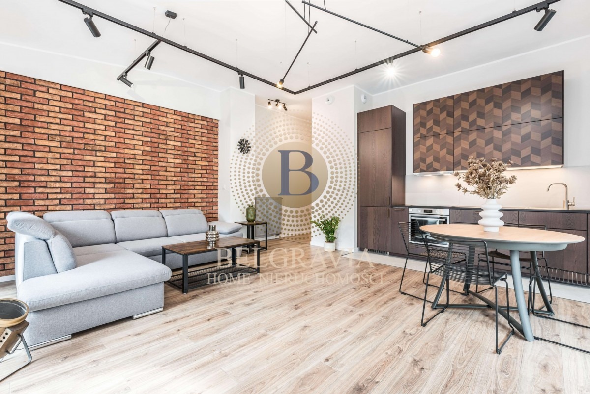 Jasne, przestronne wnętrze mieszkania z ceglaną ścianą, szarą sofą narożną, drewnianą podłogą, ciemnymi meblami kuchennymi z wzorem w jodełkę i okrągłym stołem z czarnymi krzesłami.