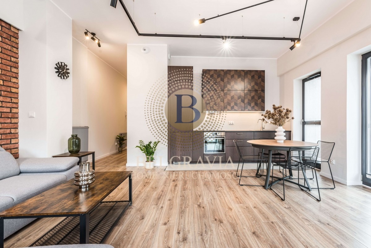 Jasne, przestronne wnętrze mieszkania z aneksem kuchennym, ceglaną ścianą i drewnianą podłogą, oświetlone reflektorami sufitowymi, z dekoracyjnymi elementami i meblami w stylu industrialnym.
