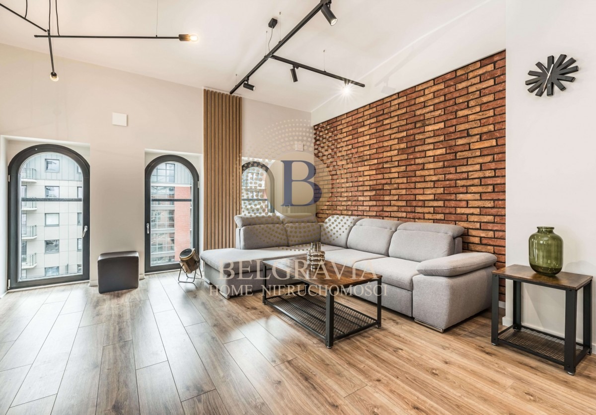 Jasny salon z szarą sofą narożną, ceglaną ścianą, drewnianą podłogą, dwoma wysokimi oknami łukowymi i czarnym oświetleniem sufitowym.