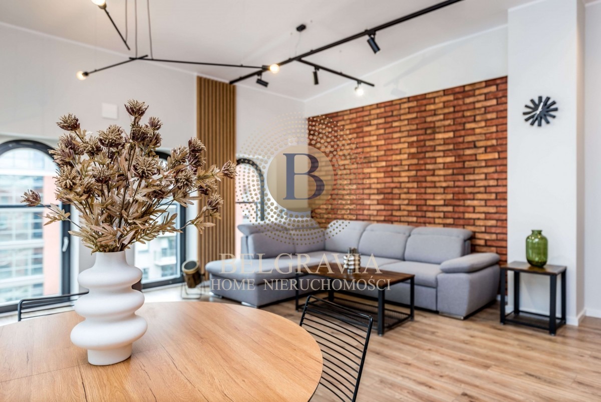 Jasne wnętrze mieszkania z ceglaną ścianą, szarą sofą narożną, drewnianym stołem i dekoracyjnym bukietem w białym wazonie na pierwszym planie.