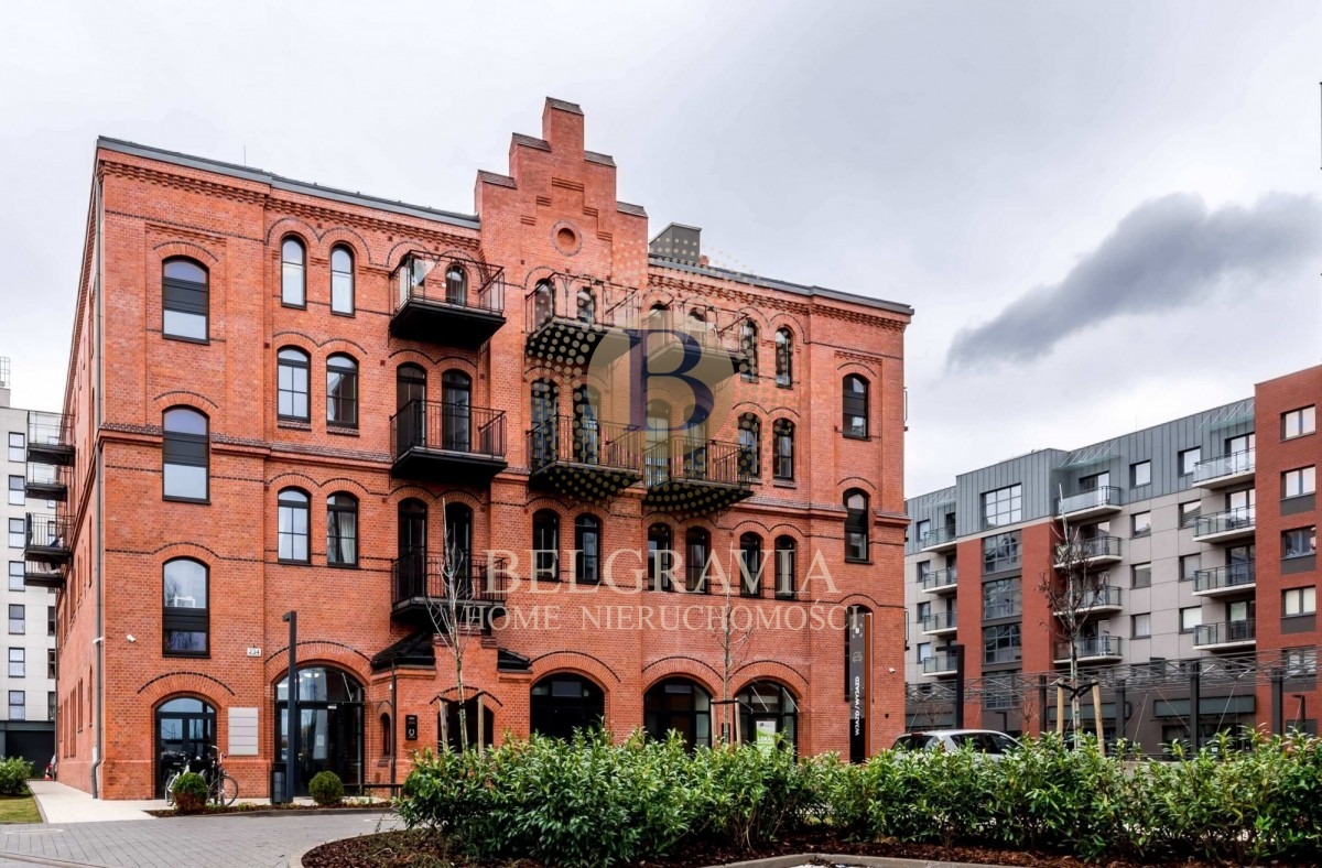 Odrestaurowany budynek z czerwonej cegły w stylu industrialnym z balkonami, widoczny z poziomu chodnika, z zielenią w pierwszym planie i nowoczesnym budynkiem w tle.