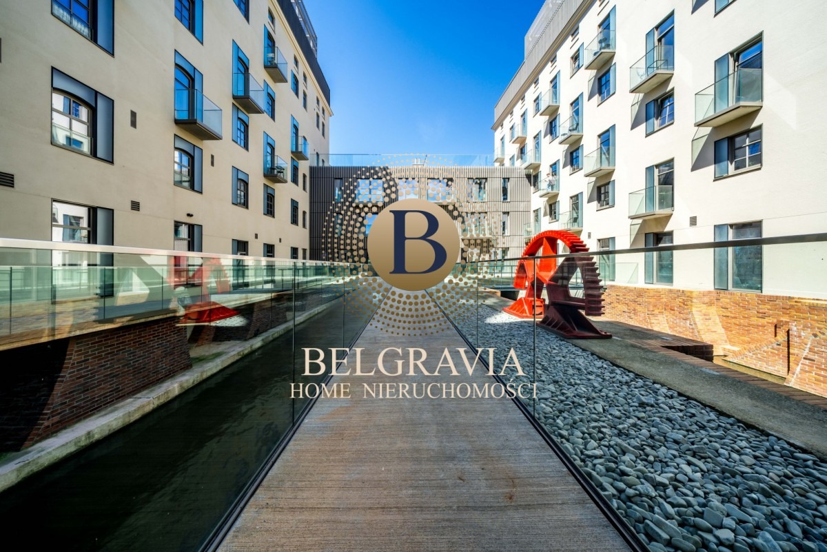 Nowoczesne budynki mieszkalne z balkonami, połączone kładką nad kanałem, z dekoracyjnym elementem w postaci czerwonego koła młyńskiego i logo firmy nieruchomościowej na pierwszym planie.