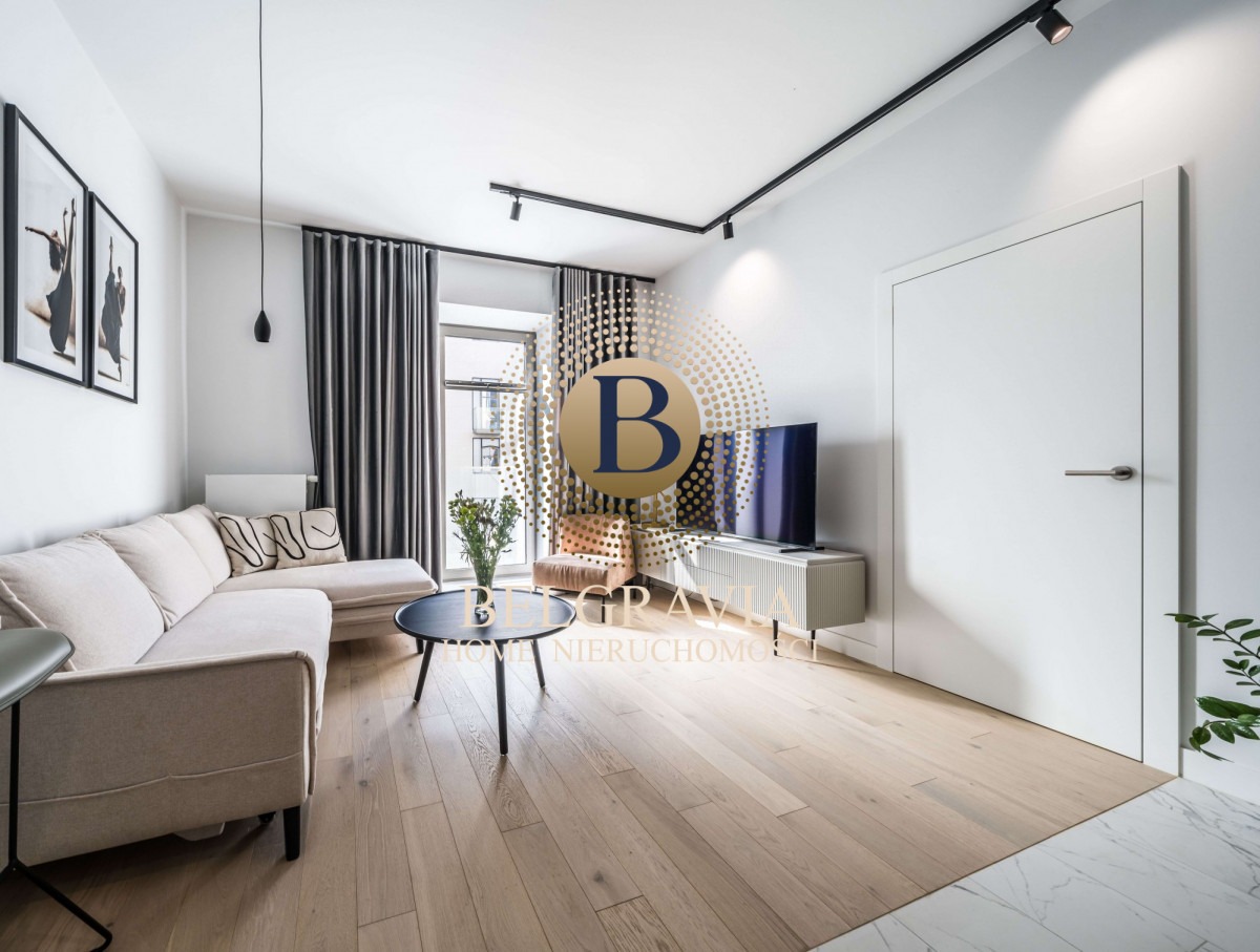 Jasny salon z narożną sofą, okrągłym stolikiem kawowym z bukietem kwiatów, telewizorem i dekoracyjnymi zasłonami; widoczne czarne szyny oświetleniowe na suficie.