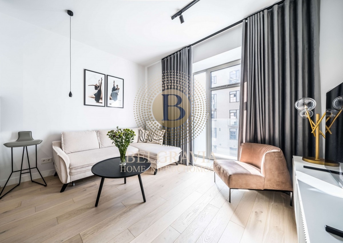 Jasne wnętrze salonu z drewnianą podłogą, beżową sofą narożną, czarnym stolikiem kawowym z bukietem kwiatów, brązowym fotelem i szarymi zasłonami przy oknie balkonowym.