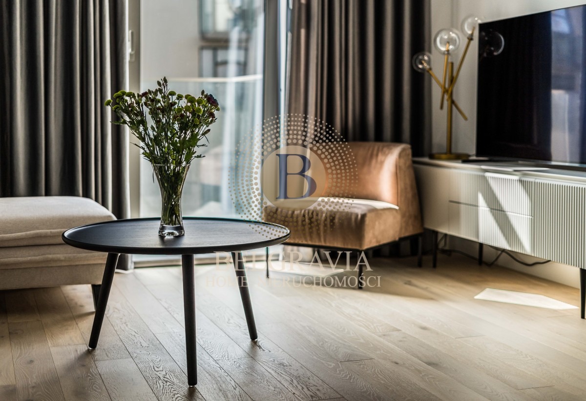 Stylowe wnętrze salonu z okrągłym czarnym stolikiem kawowym, na którym stoi wazon z kwiatami, w tle beżowy fotel, telewizor na białej szafce RTV oraz szare zasłony.