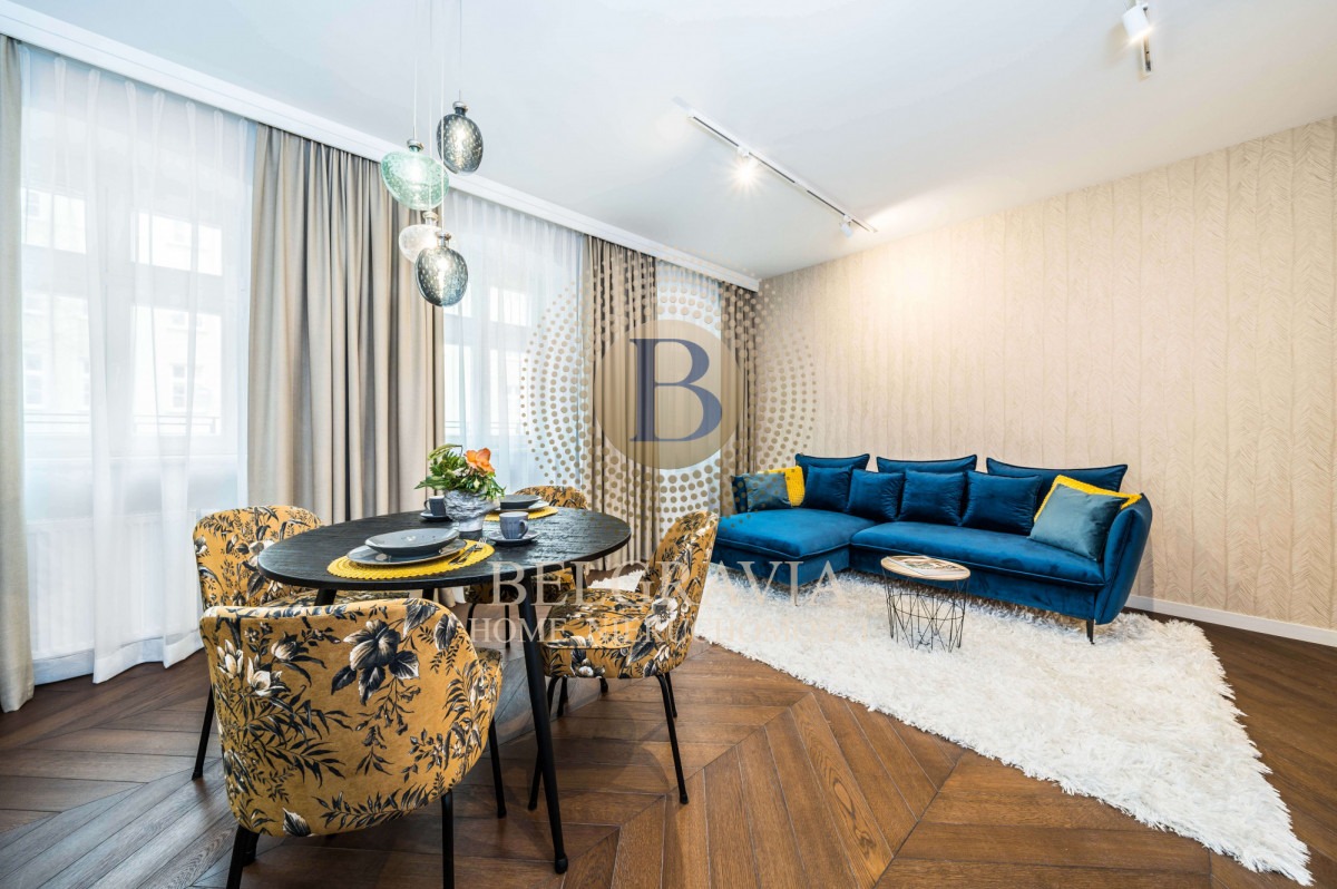 Nowoczesne wnętrze mieszkania z ciemnym okrągłym stołem, krzesłami w kwiatowy wzór, niebieską sofą narożną i parkietem ułożonym w jodełkę.