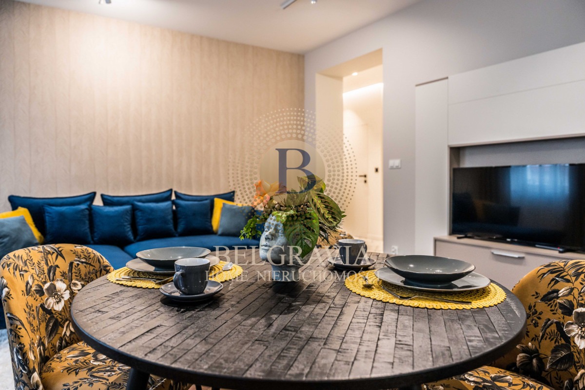 Wnętrze salonu z okrągłym stołem, nakrytym do posiłku dla dwóch osób, z widoczną sofą w odcieniach granatu i żółci oraz telewizorem w tle.