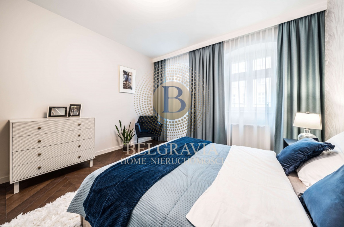 Jasna sypialnia z łóżkiem w odcieniach bieli i błękitu, komodą z dekoracyjnymi uchwytami i zasłonami w kolorze szaro-niebieskim, oświetlona naturalnym światłem z okna.