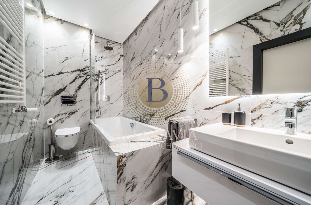 Elegancka łazienka wykończona białym marmurem z wyraźnym użyleniem, z wanną, umywalką na szafce, toaletą i grzejnikiem drabinkowym, oświetlona wiszącymi lampami i z lustrem z podświetleniem LED.