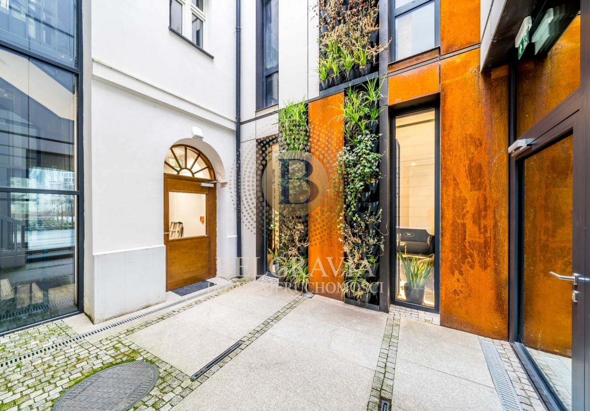 Dziedziniec budynku z pionowymi ogrodami i elementami z oksydowanej stali, widoczne drzwi wejściowe i okna.