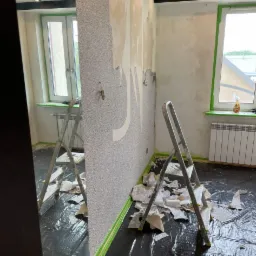 Pokój w trakcie usuwania starej tapety z fragmentami gładzi, zabezpieczony folią i taśmą malarską, dwie drabiny aluminiowe, widoczne punkty oświetleniowe na suficie.