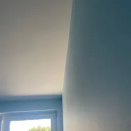 Świeżo pomalowane, błękitne ściany w pokoju z widokiem na sąsiednie budynki przez okno; sufit w jasnym odcieniu bieli.