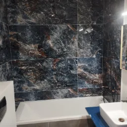 Nowoczesna łazienka z płytkami imitującymi marmur w odcieniach granatu i brązu, białą wanną i umywalką nablatową z czarną armaturą, oświetleniem LED i złotymi detalami.