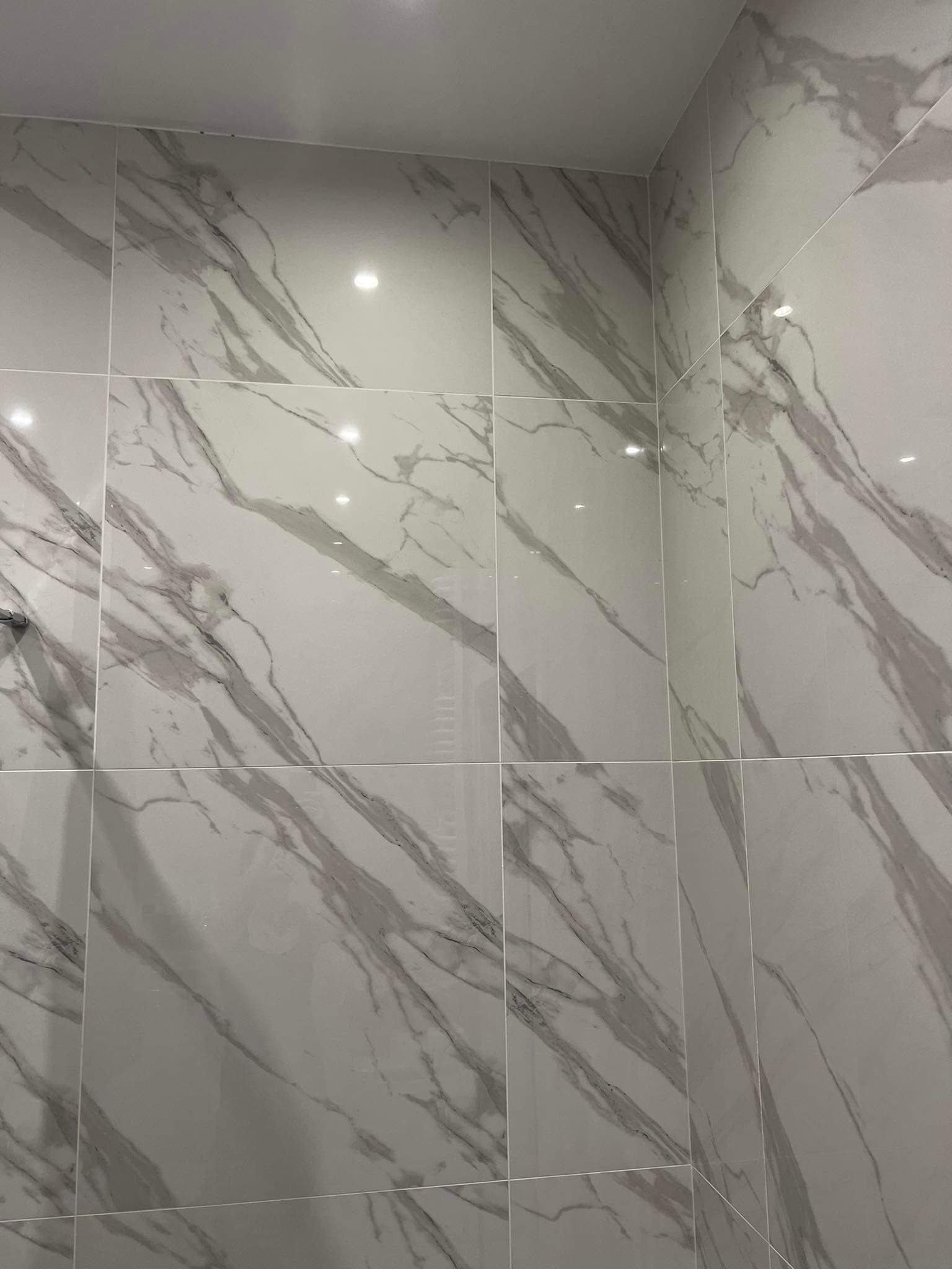 Ściana łazienki wyłożona dużymi, kwadratowymi płytkami imitującymi biały marmur z szarymi żyłami, widoczne fugi i odbicia światła.