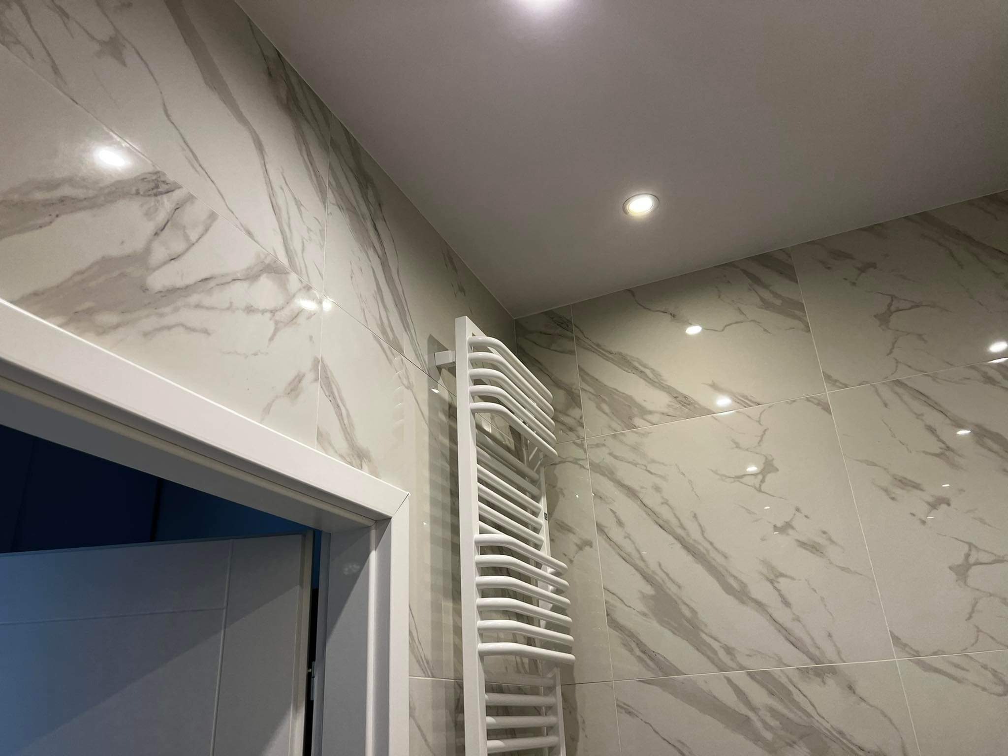 Nowoczesna łazienka z białymi płytkami imitującymi marmur, grzejnik drabinkowy w kolorze białym, oświetlenie punktowe w suficie i widoczne fragmenty białych drzwi.