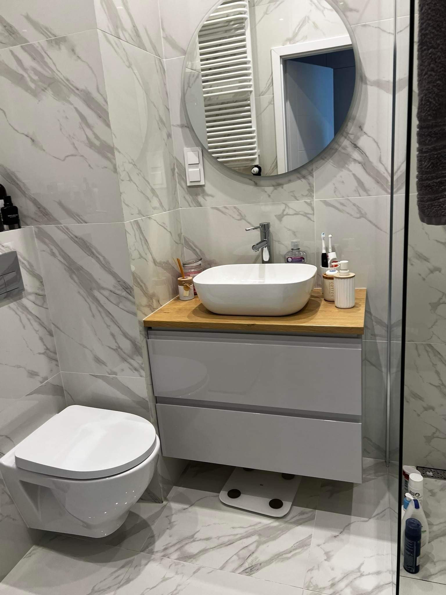 Nowoczesna łazienka z marmurowymi płytkami, wiszącą toaletą, okrągłym lustrem z podświetleniem LED, białą umywalką nablatową na drewnianym blacie i szarą szafką pod umywalką.