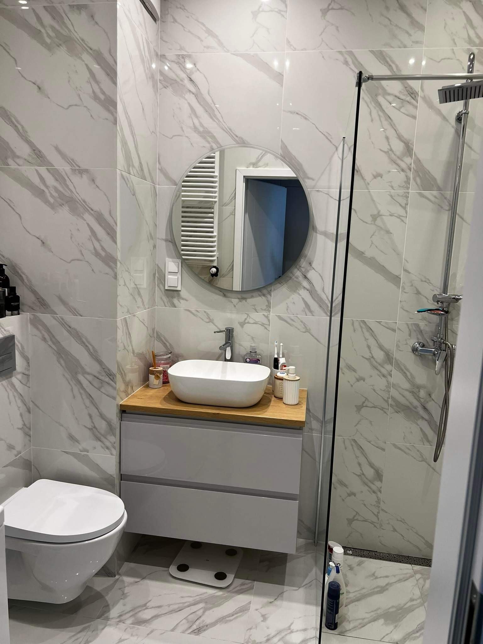 Nowoczesna łazienka z biało-szarymi płytkami imitującymi marmur, okrągłe lustro z widocznym grzejnikiem drabinkowym w odbiciu, biała umywalka nablatowa na szarej szafce z drewnianym blatem...