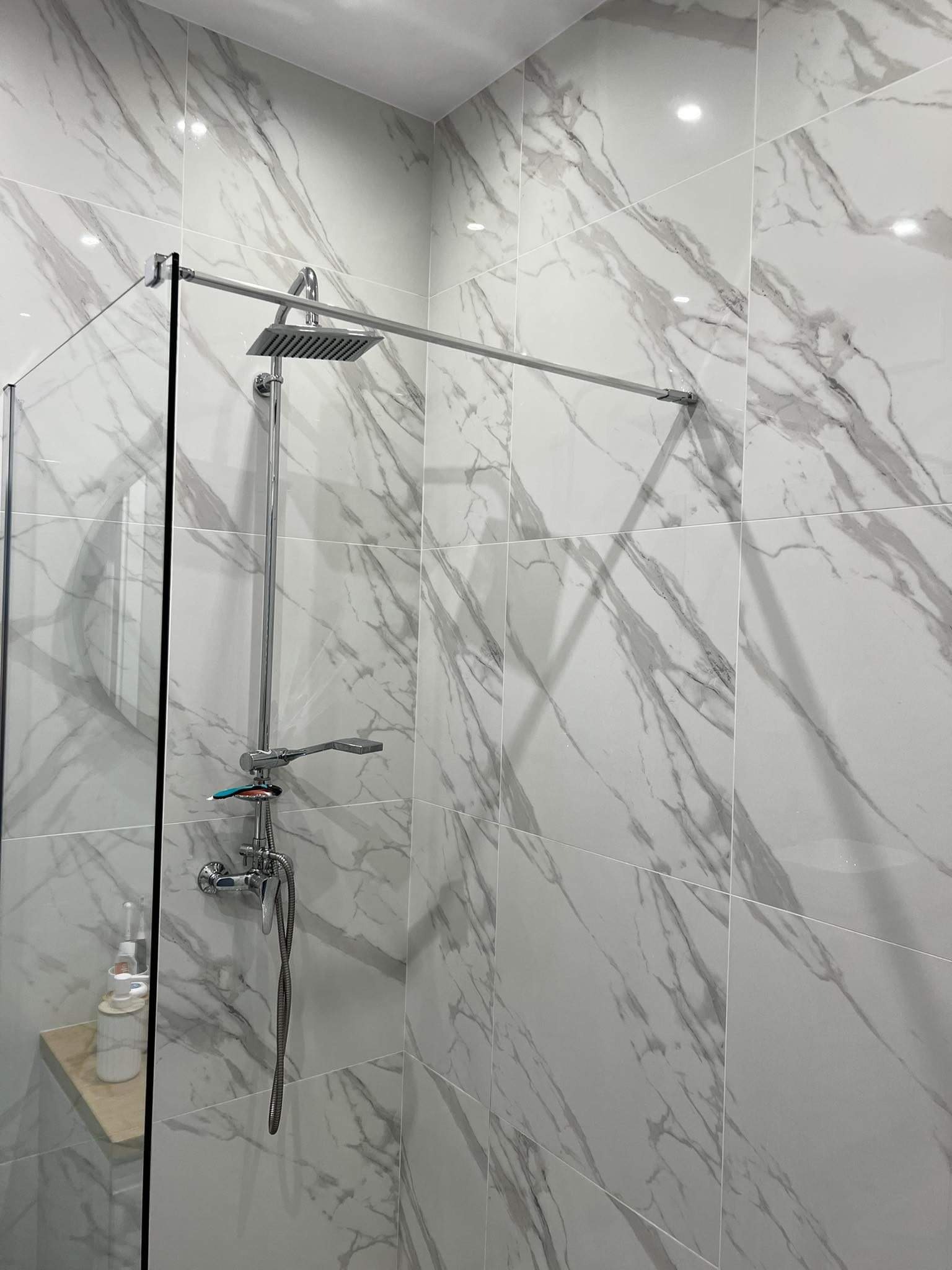 Nowoczesna kabina prysznicowa ze szklaną ścianką i deszczownicą, wykończona białymi płytkami imitującymi marmur, z widocznymi kosmetykami na półce.