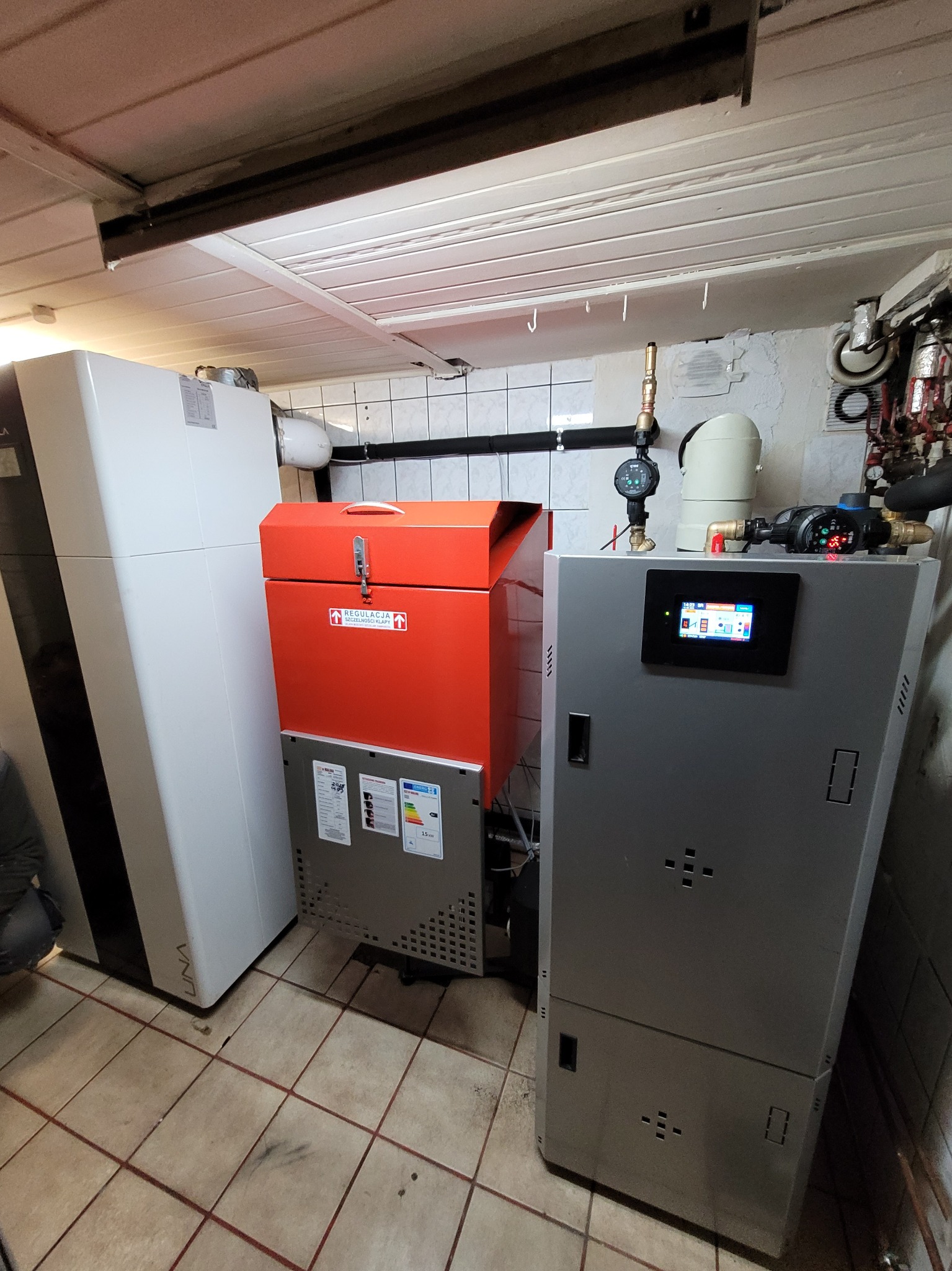 Nowoczesna kotłownia z piecem na paliwo stałe w kolorze pomarańczowym, pompą ciepła i szarym kotłem z panelem sterowania, zainstalowana w pomieszczeniu z kafelkami na ścianach i podłodze.