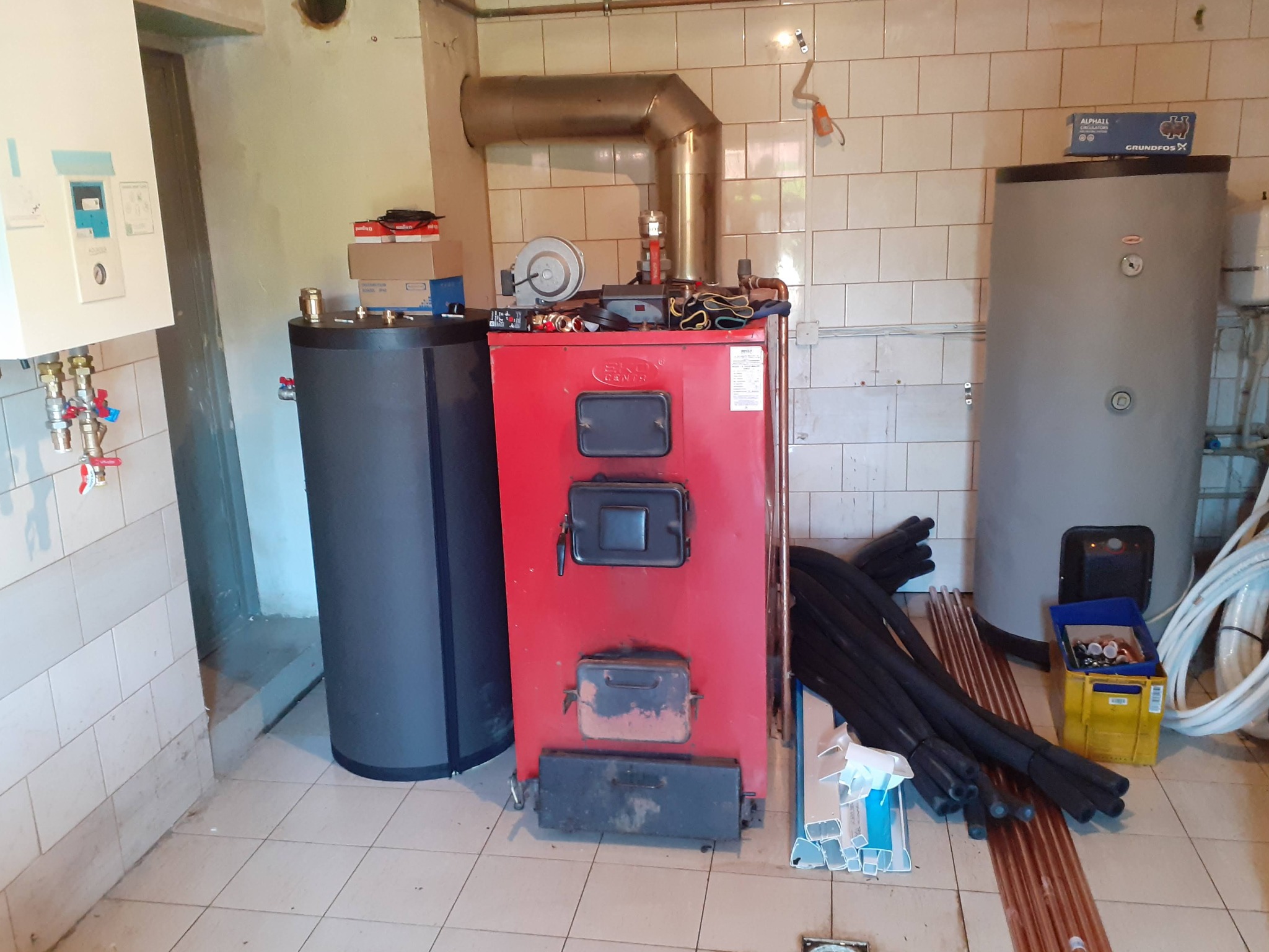 Czerwony piec na paliwo stałe z podłączonym kominem, obok czarny zbiornik akumulacyjny i szary zasobnik ciepłej wody użytkowej. Na podłodze rury izolowane i miedziane. W tle białe kafelki.