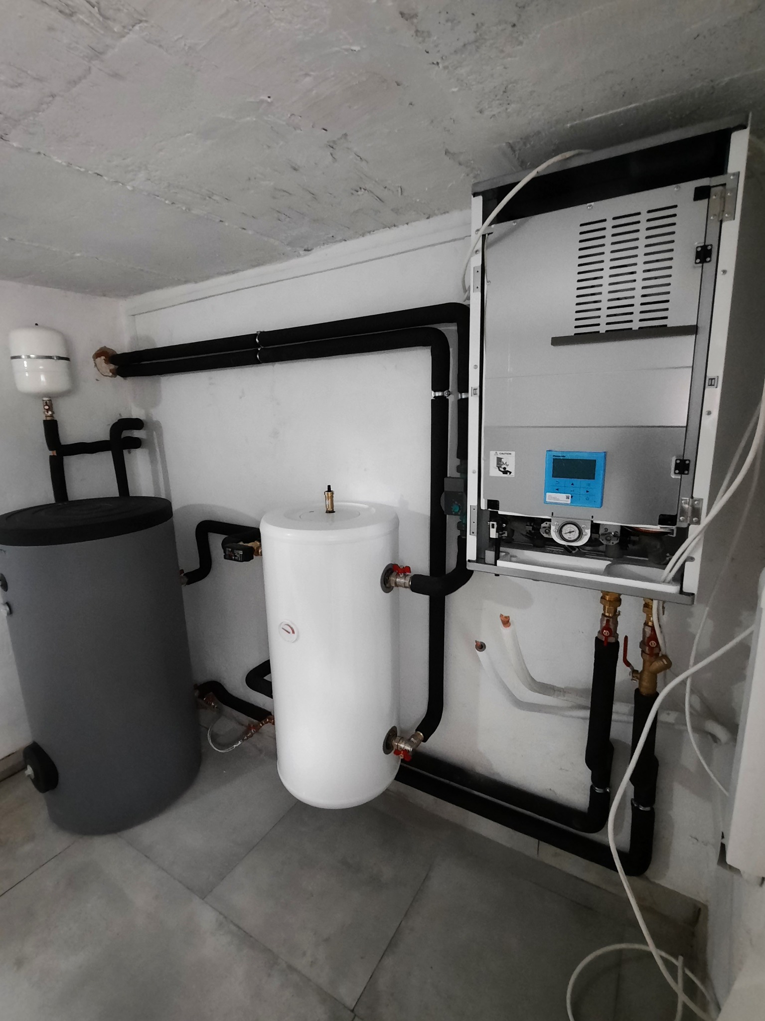 Zainstalowany, nowoczesny piec gazowy kondensacyjny z dwoma zbiornikami buforowymi w kotłowni z szarą podłogą i surowym betonowym sufitem.