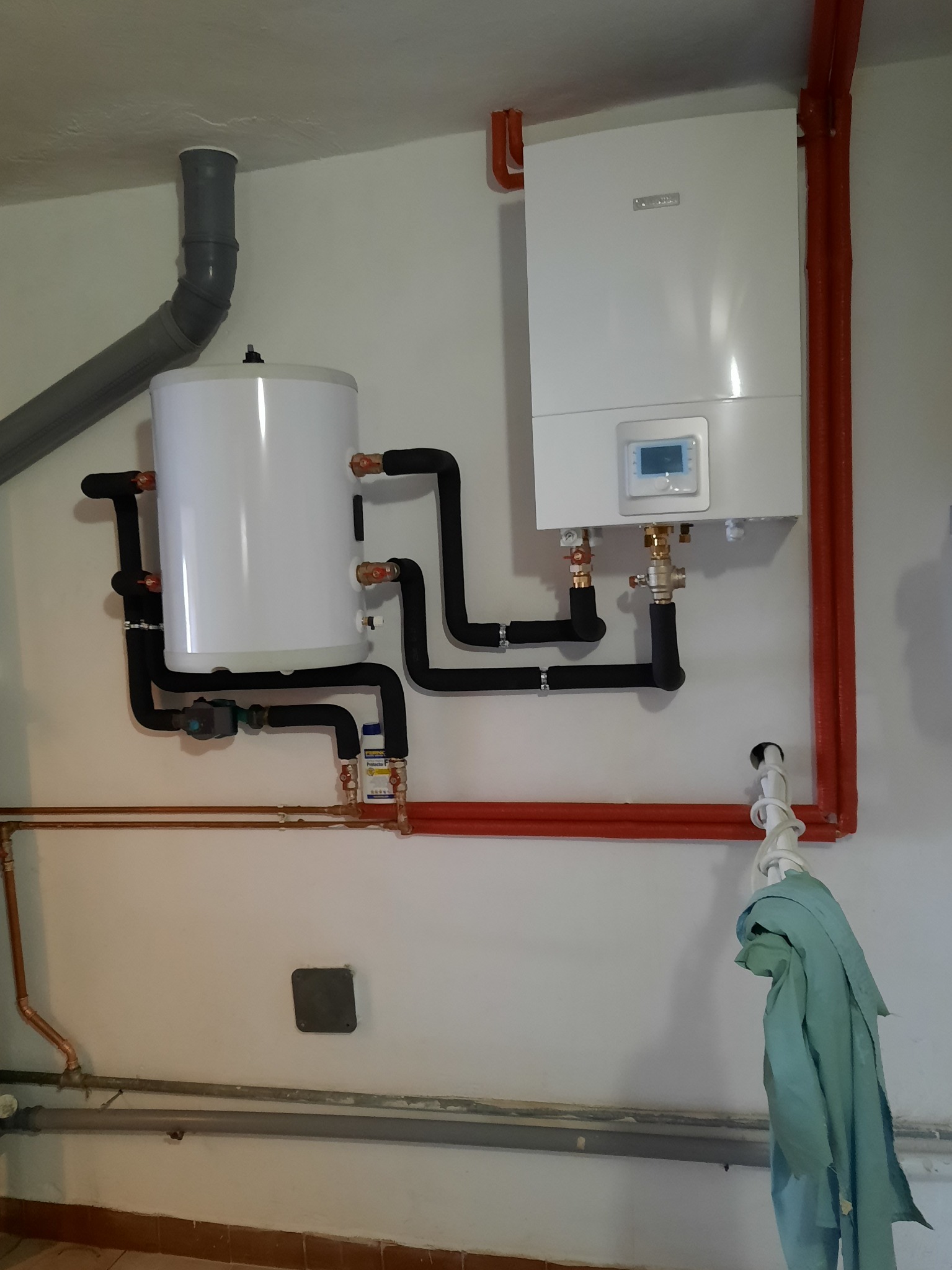 Biały, wiszący na ścianie piec gazowy z podłączonym zasobnikiem wody, rury miedziane i czarne izolowane, szary przewód wentylacyjny, fragment ściany i podłogi.