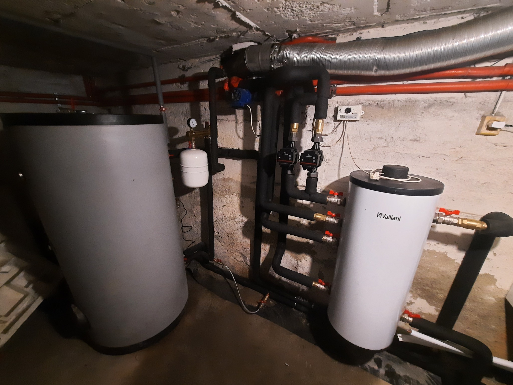 Dwa zbiorniki akumulacyjne, szary i biały marki Vaillant, połączone rurami w izolacji, z widocznymi zaworami i pompami obiegowymi, zainstalowane w piwnicy z surowym betonowym sufitem i ścianami...