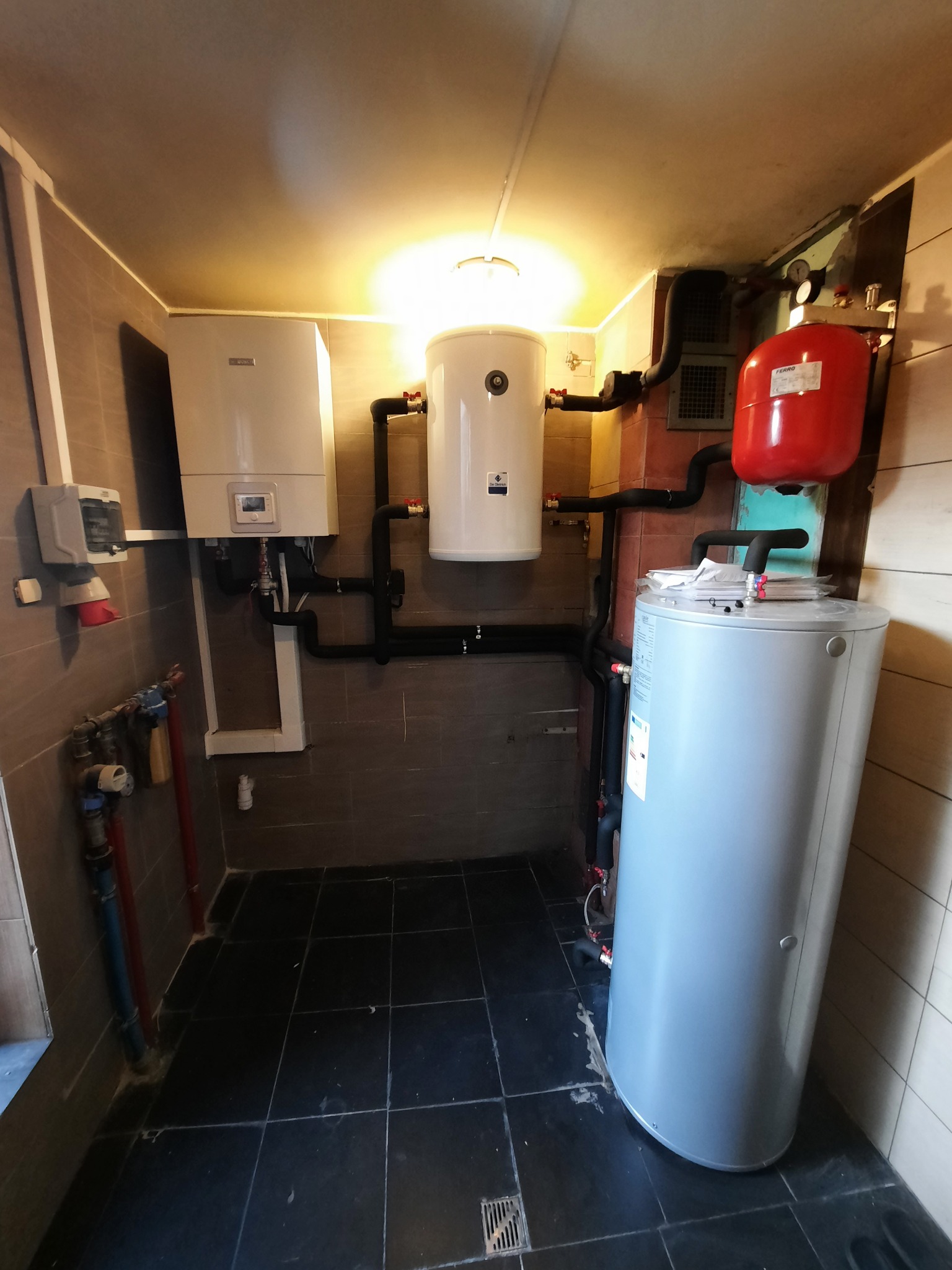 Kompaktowa kotłownia w pomieszczeniu wyłożonym brązowymi i szarymi płytkami, z wiszącym białym kotłem, zasobnikiem ciepłej wody i czerwonym zbiornikiem wyrównawczym, połączonymi czarnymi rurami.