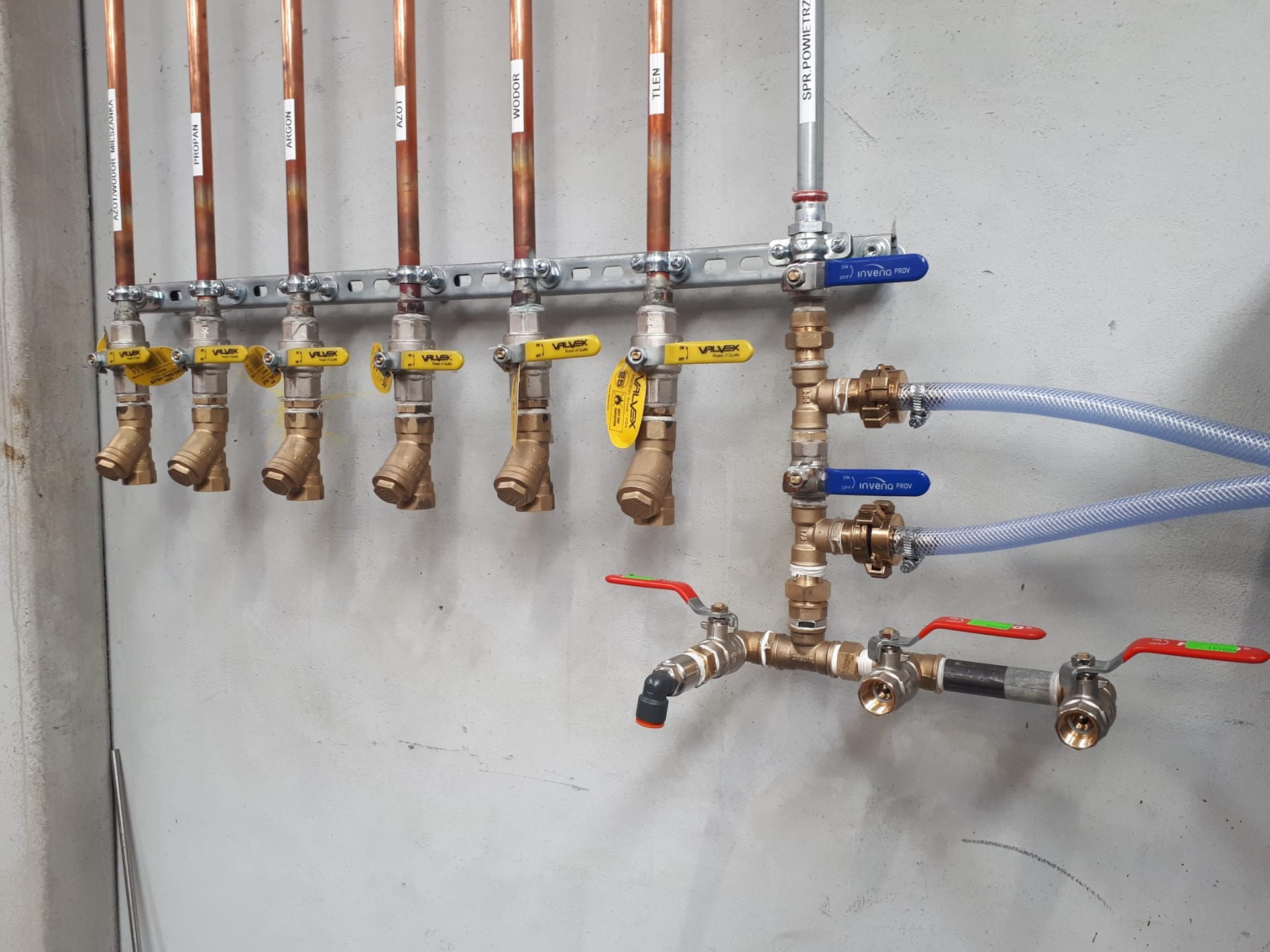 Rozbudowana instalacja gazowa z miedzianymi rurami i zaworami oznaczonymi nazwami gazów (azot/wodór, propan, argon, azot, wodór, tlen, sprężone powietrze), zamontowana na szarej, surowej ścianie.
