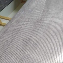 Fragment szarego, prążkowanego dywanu z widocznymi śladami użytkowania i fragmentem konstrukcji meblowej w tle.
