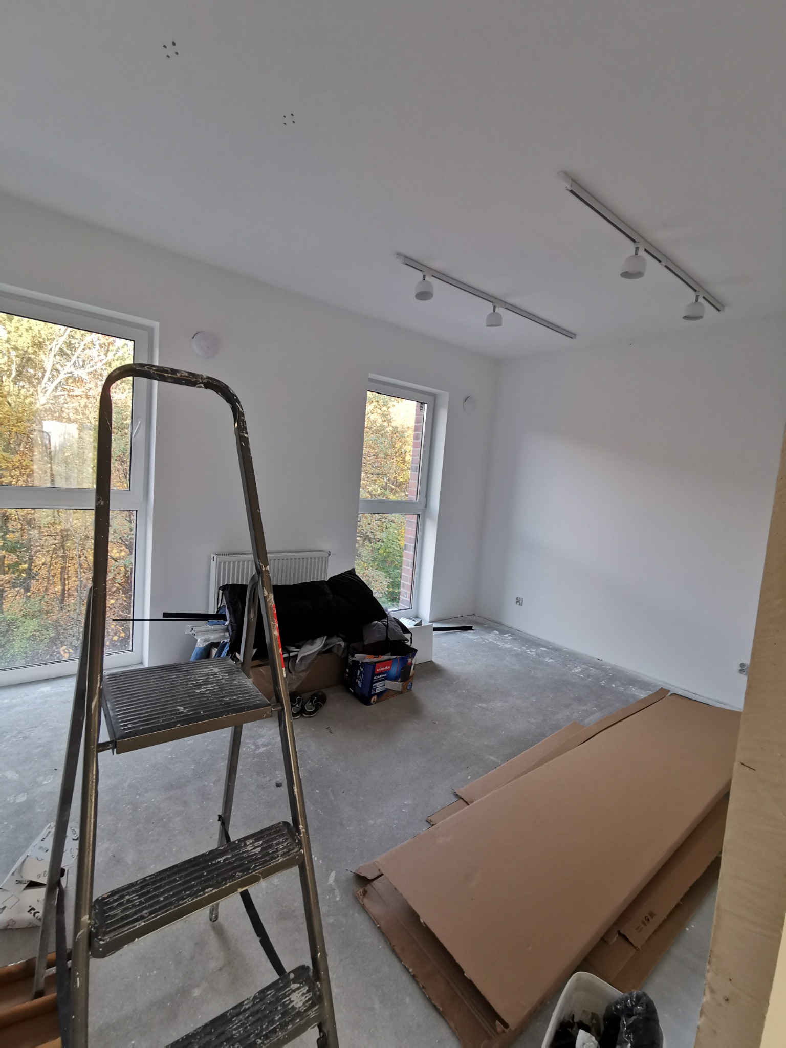 Pokój w trakcie prac wykończeniowych: świeżo pomalowane ściany na biało, metalowa drabina na pierwszym planie, dwa okna z widokiem na drzewa, kartony na podłodze i zamontowane oświetlenie szynowe.