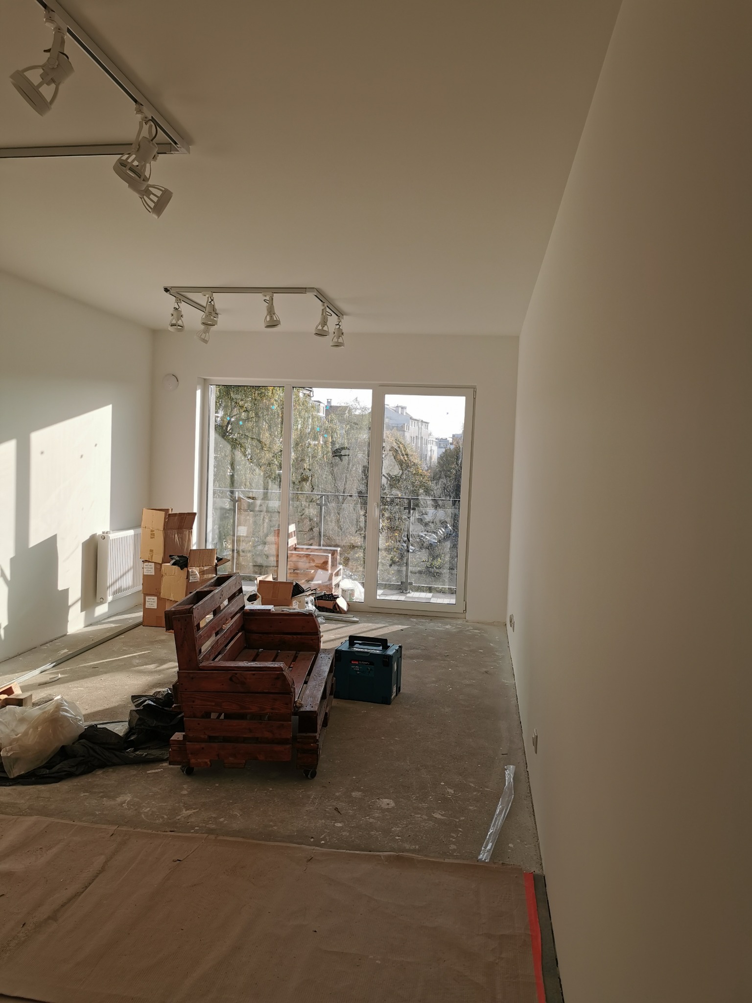 Wnętrze pokoju w trakcie remontu: świeżo pomalowane ściany, zabezpieczona podłoga, widoczne kartony, drewniana sofa z palet i narzędzia, okno balkonowe z widokiem na zewnątrz.