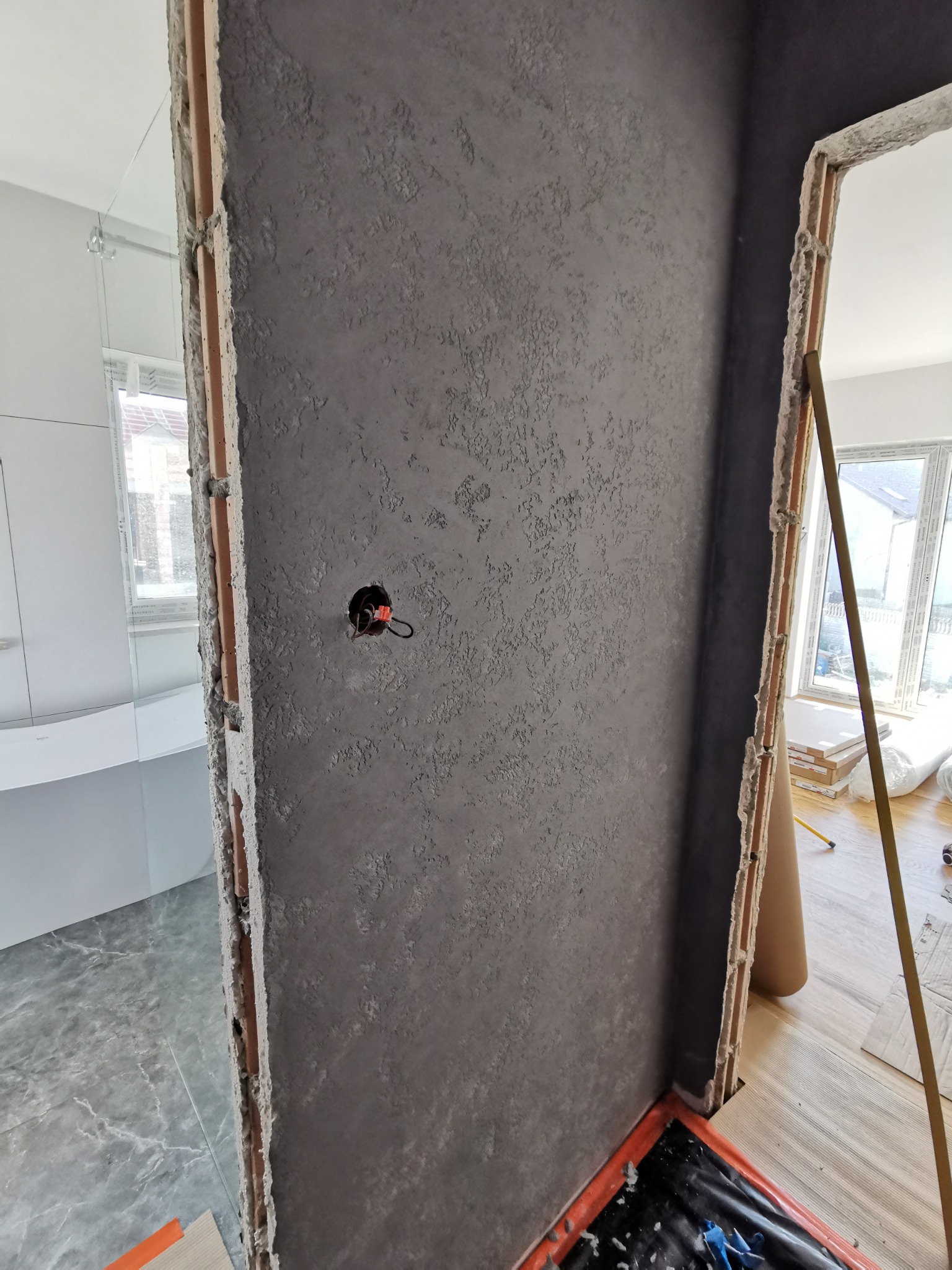 Ściana pokryta szarą, strukturalną gładzią w trakcie prac wykończeniowych, widoczne niedoskonałości obróbki ościeżnic i wystający kabel elektryczny ze ściany, fragment wykończonej łazienki w tle.