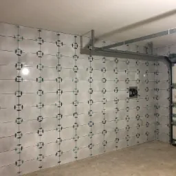 Świeżo ułożone, jasnoszare płytki na ścianie garażu, z użyciem krzyżyków dystansowych, widoczne metalowe elementy konstrukcyjne nad drzwiami garażowymi.