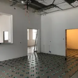 Przygotowanie podłoża pod płytki w dużej przestrzeni biurowej, widoczne poziomice i krzyżyki dystansowe, surowy stan ścian i sufitu z widoczną instalacją wentylacyjną.