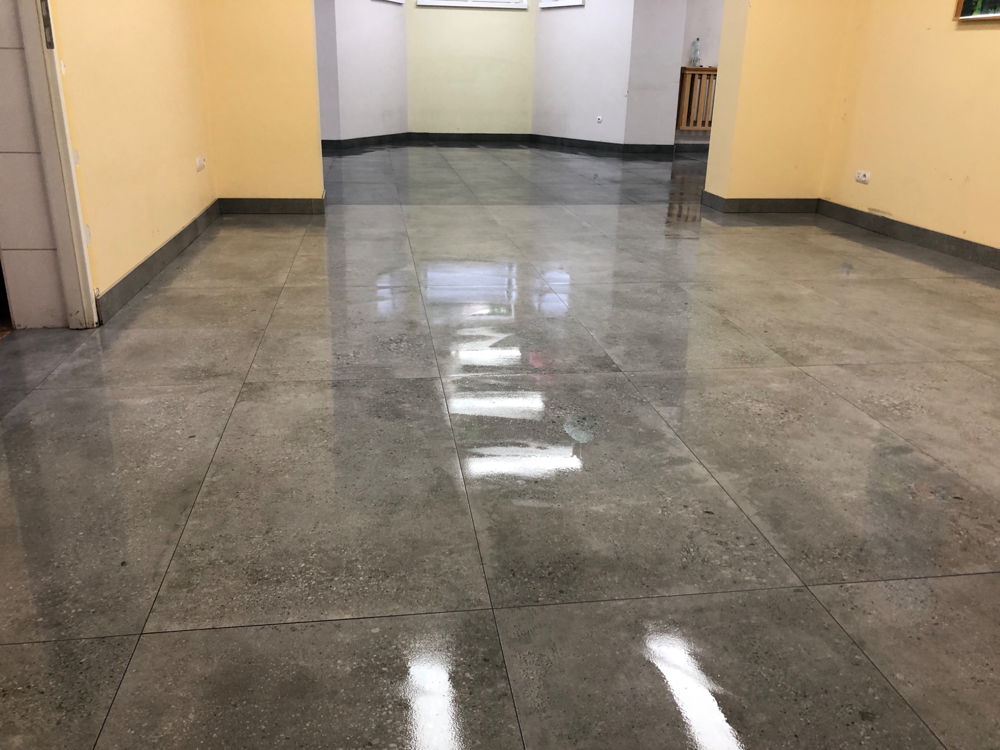 Polerowana, szara posadzka z płytek w pustym pomieszczeniu z żółtymi ścianami i widocznymi gniazdkami elektrycznymi.