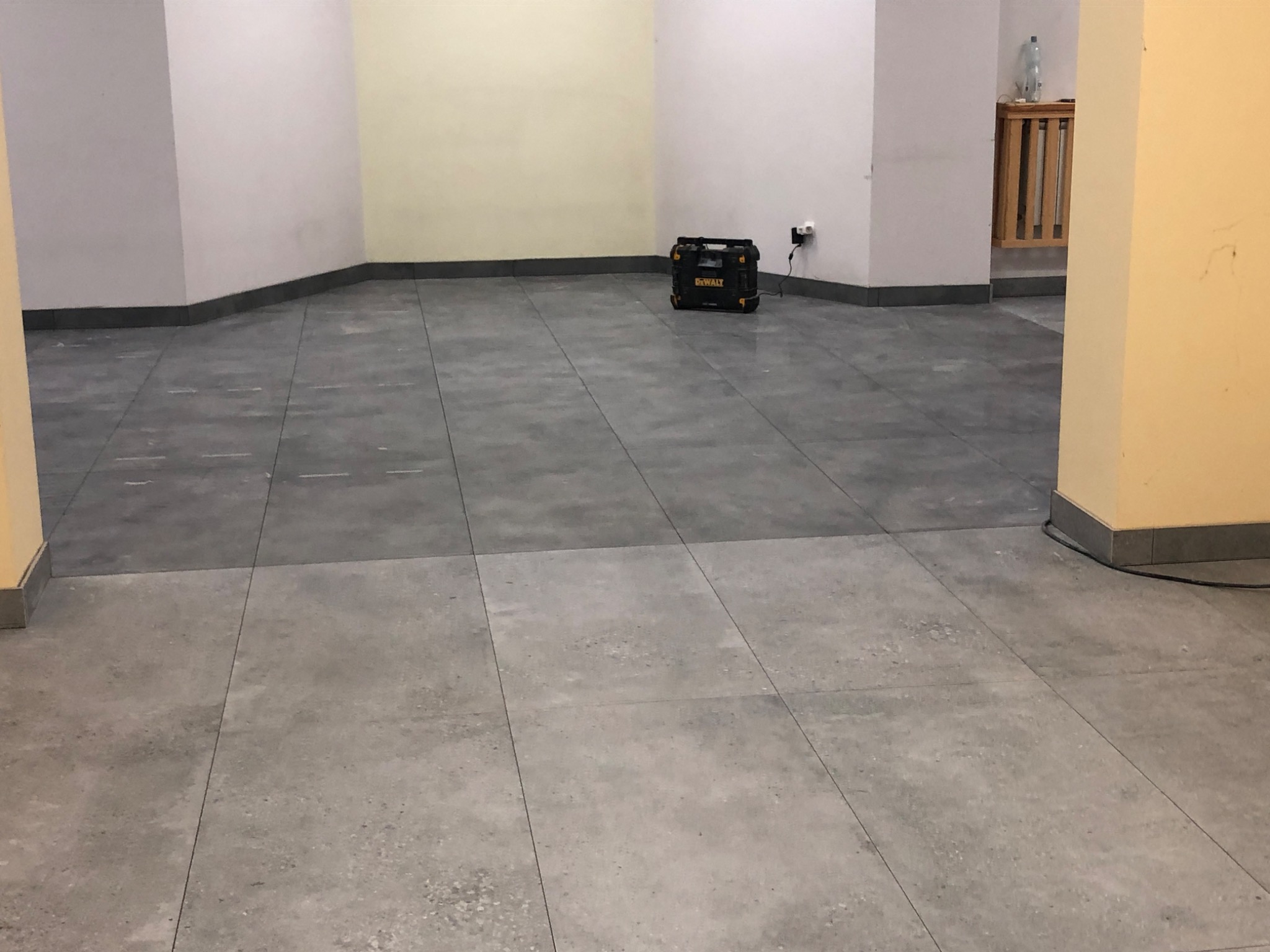 Wykończona podłoga z szarych, matowych płytek w pustym pomieszczeniu, widoczny czarny agregat prądotwórczy DeWalt przy ścianie.
